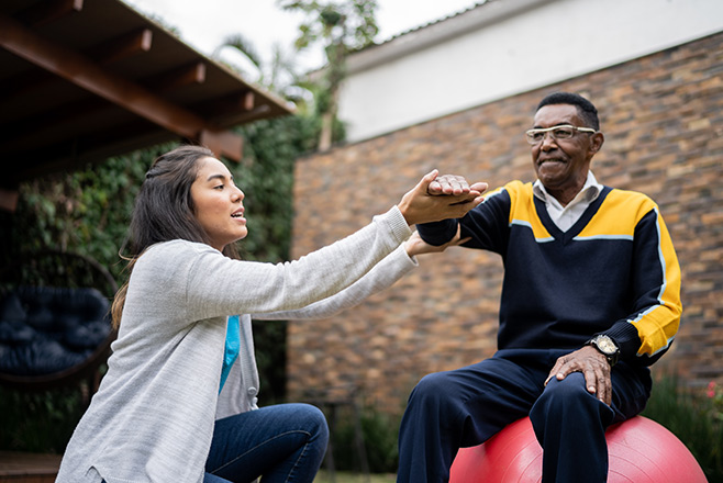 Woman holding a mans arm whilst he sits on an exercise ball
