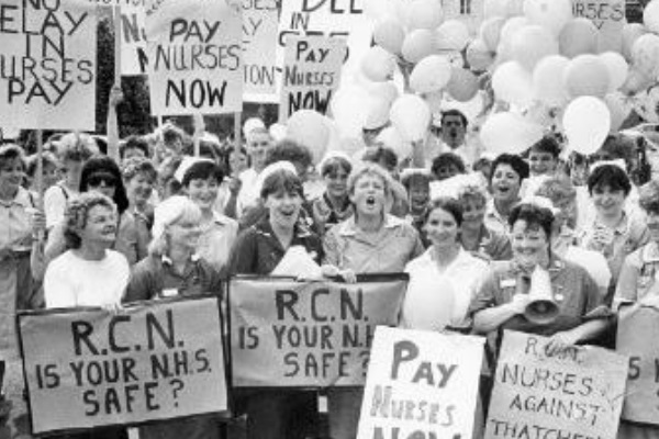 A black and white photo of an RCN demonstration with nursing staff holding signs saying 'Pay nurses now' and 'RCN - Is your NHS safe?'