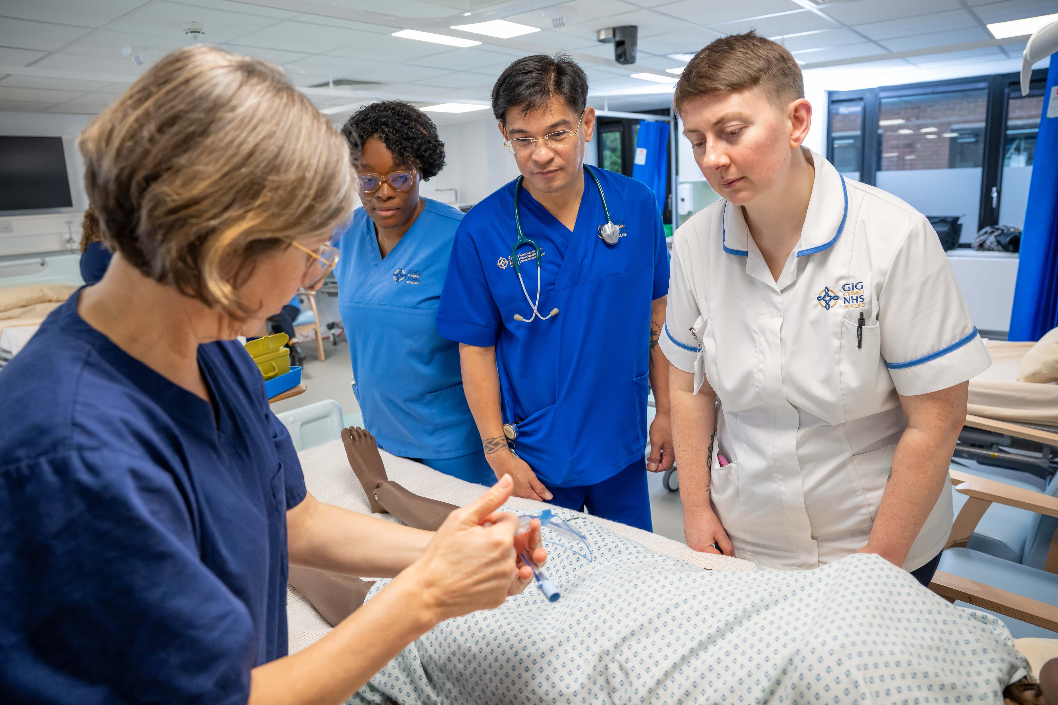 Nurses are standing over a a practice table. One nurse is demonstrating with an iv tube, while the other three look on.