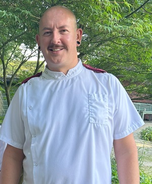 Garritt Brown is standing in garden, he is wearing his white nursing uniform and smiling broadly
