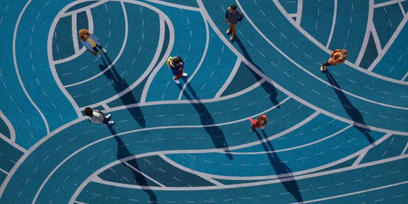 Pathways with people pictured from a bird's eye view (from above) with long shadows, going along different intersecting blue roads leading out of the picture and going around in swirls and curves