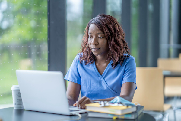 Black woman in nursing scrubs sits at a laptop computer to study