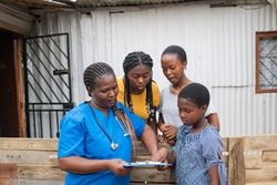 A healthcare worker in blue scrubs with a stethoscope around her neck shows a clipboard to a group of three children and teenagers, who are gathered around her, attentively looking at the document.