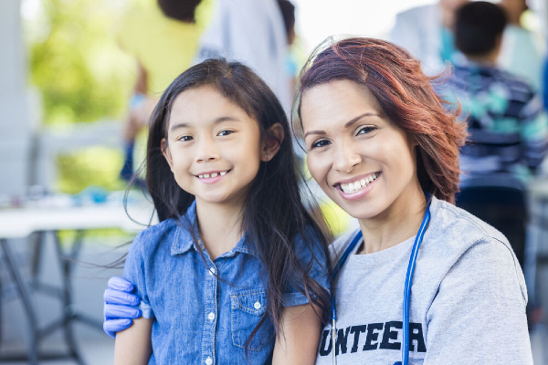 A smiling female volunteer wearing a stethoscope poses with a young girl in a casual outdoor setting, possibly at a community or health outreach event.