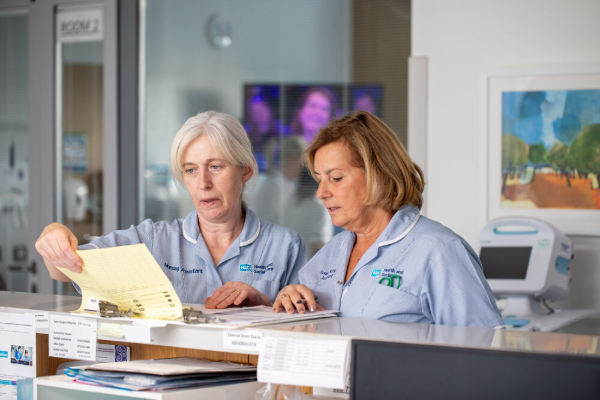 Two nurses in light blue uniforms review paperwork together at a hospital reception desk.