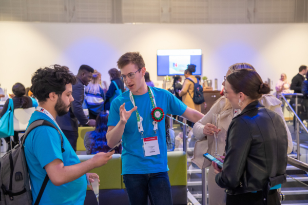 A group of young people talking and looking at smartphones during a networking event or conference in a brightly lit indoor space.