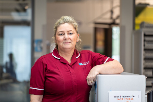A woman in a red NHS Health and Social Care uniform stands in a reception area, looking at the camera.