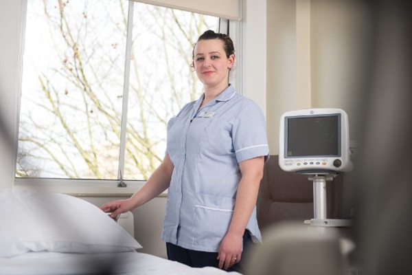 A nursing support worker standing next to a hospital bed and smiling