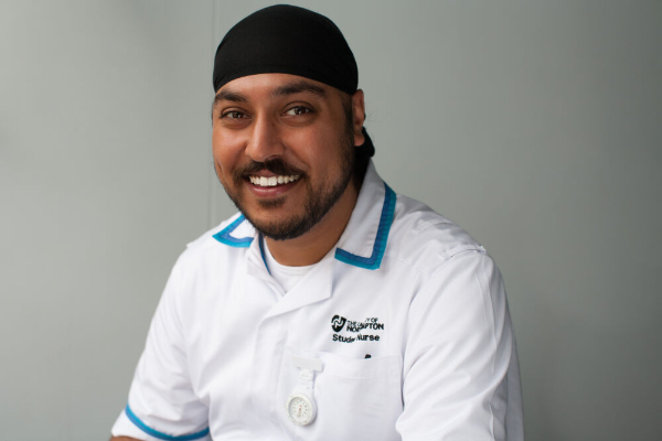A male nurse in a white uniform with blue accents, smiling warmly against a plain grey background.