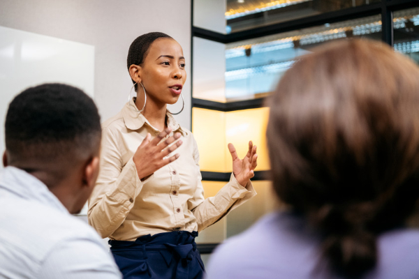 A woman in a beige blouse speaking to a small group in a modern office setting, using hand gestures to emphasize her points.