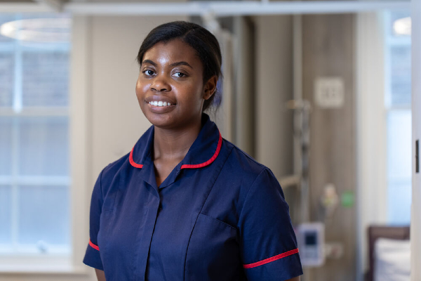 A female nurse in a navy blue uniform with red trim, standing in a hospital setting with a confident smile.
