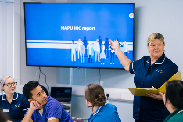 A nurse presenting at a screen to an audience of four other nurses. The screen reads 'HAPU IHC report'.