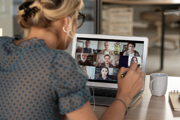 A person viewed from over the shoulder participates in a video conference call on a laptop, with multiple attendees visible in a grid on the screen.