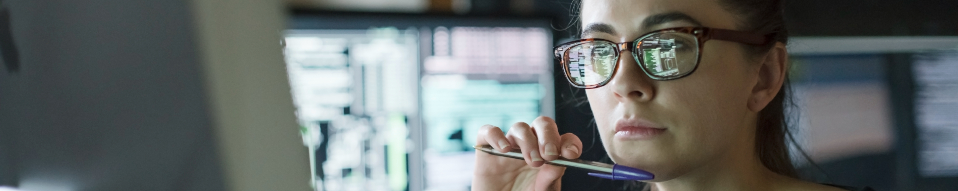 A focused woman wearing glasses looks at a computer monitor, with code and data reflected in her lenses.