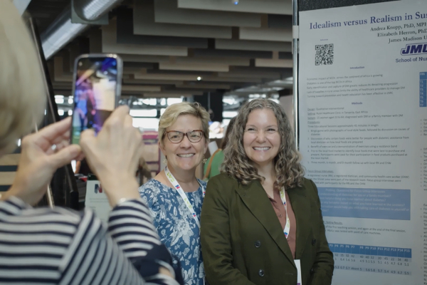 A person takes a smartphone photo of two people smiling as they stand in front of a scientific research poster at a conference.
