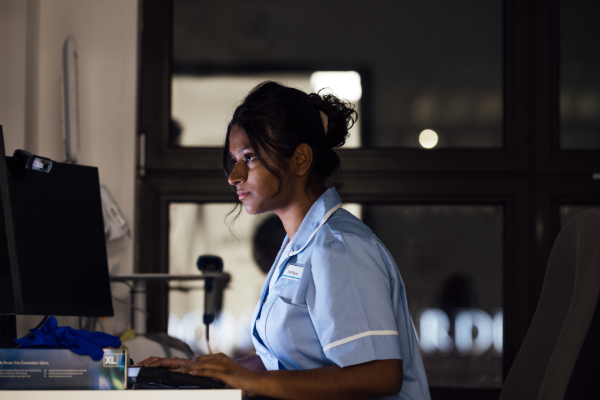 A nurse in a light blue uniform sits at a desk and works on a computer at night.