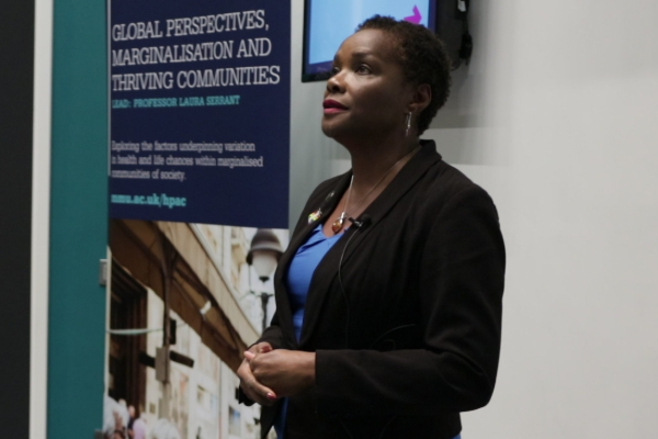 A person in a black blazer speaks at a conference or lecture, standing next to a banner titled "Global Perspectives, Marginalisation and Thriving Communities."