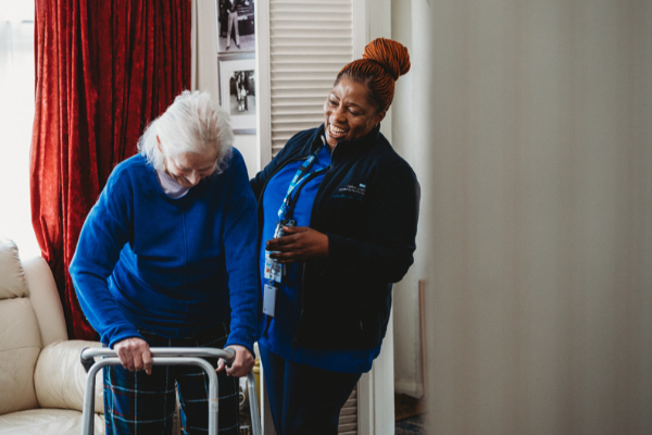 A female nurse smiling and providing support to an elderly person who is using a walking frame indoors.