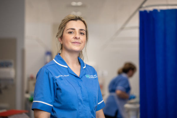 A female nurse in a blue uniform standing in a clinical setting, such as a hospital ward or clinic.