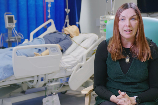 Picture of a presenter talking to camera in front of a hospital bed