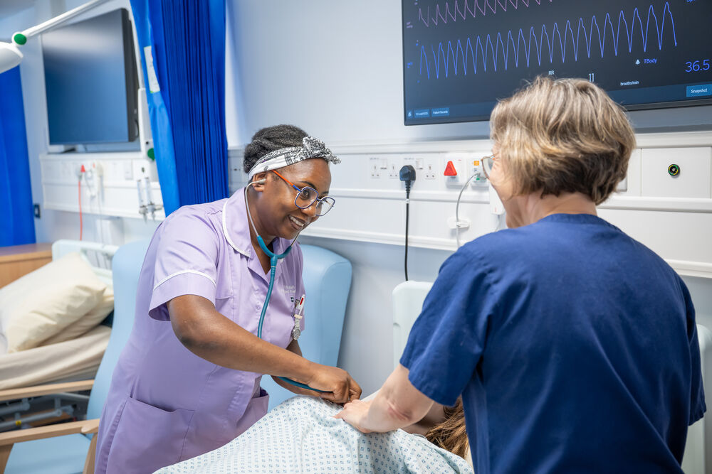 A nursing support worker standing over a hospital bed and smiling at a colleague