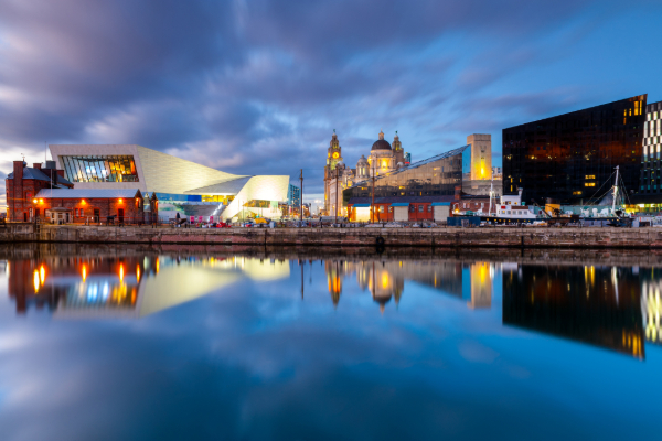 A scenic waterfront cityscape at twilight featuring illuminated modern buildings reflecting in the calm water.