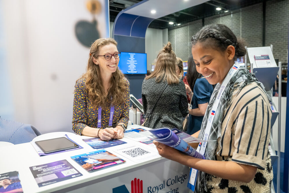 Two women smiling and conversing at an exhibition stand; one stands behind the counter while the other reviews a brochure she is holding.