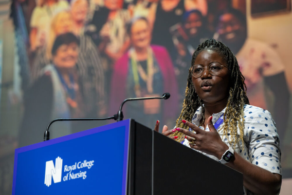 A woman speaking at a podium with a Royal College of Nursing logo, gesturing with her hands during a presentation.