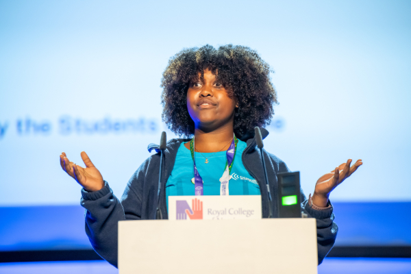 A speaker standing behind a podium with hands raised in an open gesture while addressing a crowd.