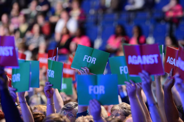 A large audience in a conference hall raising green "FOR" and red "AGAINST" voting cards.