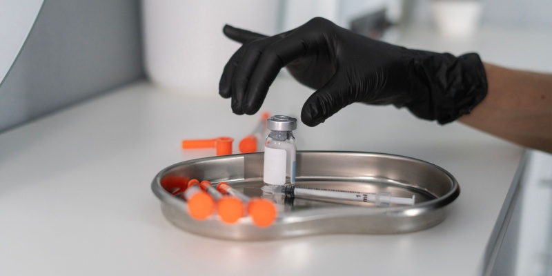 a hand with a black glove reaches for a bottle of botox, beside a syringe