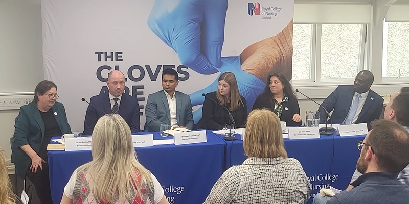 Six politicians from leading parties sit beside each other on a panel, the backs of some audience members is visible