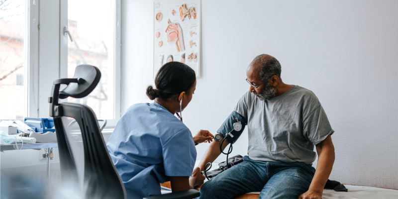 a man sitting on a doctor's couch with a woman in a light blue uniform taking his blood pressure