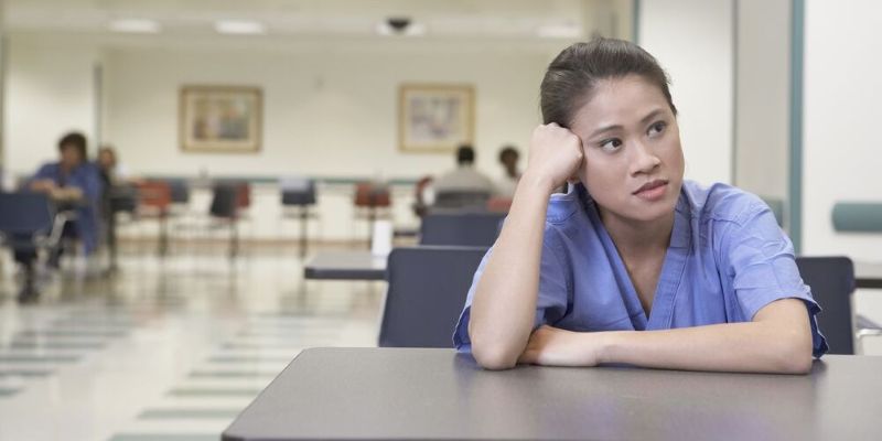 A photo of a woman wearing blue scrubs sat at a table with her arms leant on the table. The woman looks pensive. 