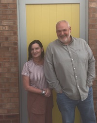 Two staff members stood in front of a yellow door looking happy at PIPs.