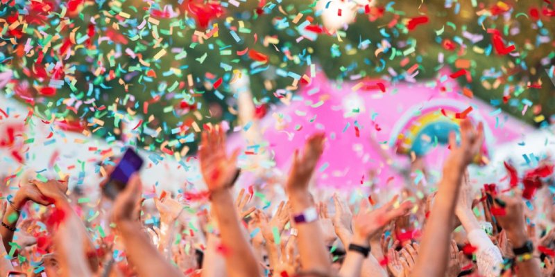 A crowd of hands in the air with colourful blue, red and yellow confetti raining down on them, and a pink umbrella and a little rainbow shape held aloft in the background, in front of green shapes at the top of the image, presumably trees further in the background.