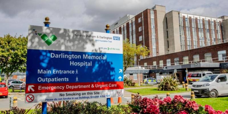 Darlington Memorial Hospital sign with pink and green foliage underneath it, slightly to the left of the picture, in front of the hospital blocks (one storey and multi-storey) which take up the right hand side of the picture
