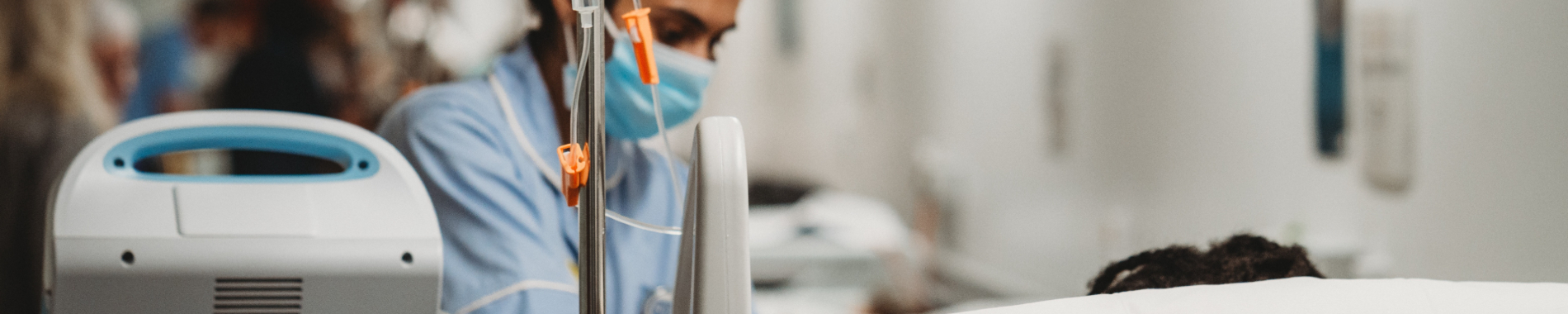 A nurse in a light blue uniform and surgical mask tends to an IV drip for a patient in a hospital bed, with the background softly blurred.