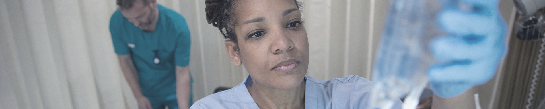 An image of a female nurse holding an IV drip, with a male nurse working in the background.