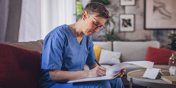 Female nurse doing paperwork Older female nurse in scrubs sat on the sofa doing paperwork