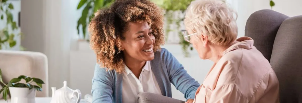A smiling carer sits and has a warm, friendly conversation with an older woman in her home.
