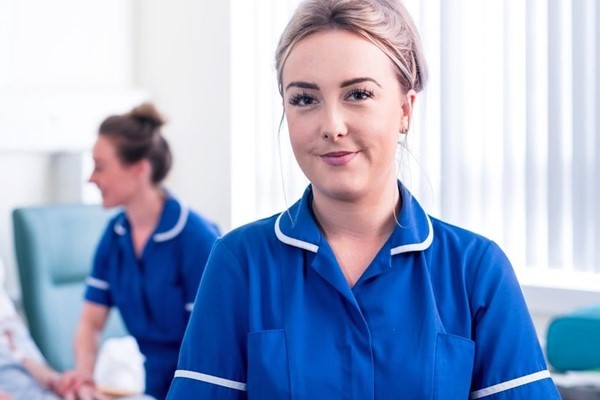 A nurse in a blue uniform looks confidently at the camera, with a colleague attending to a patient in the blurred background of a hospital ward.