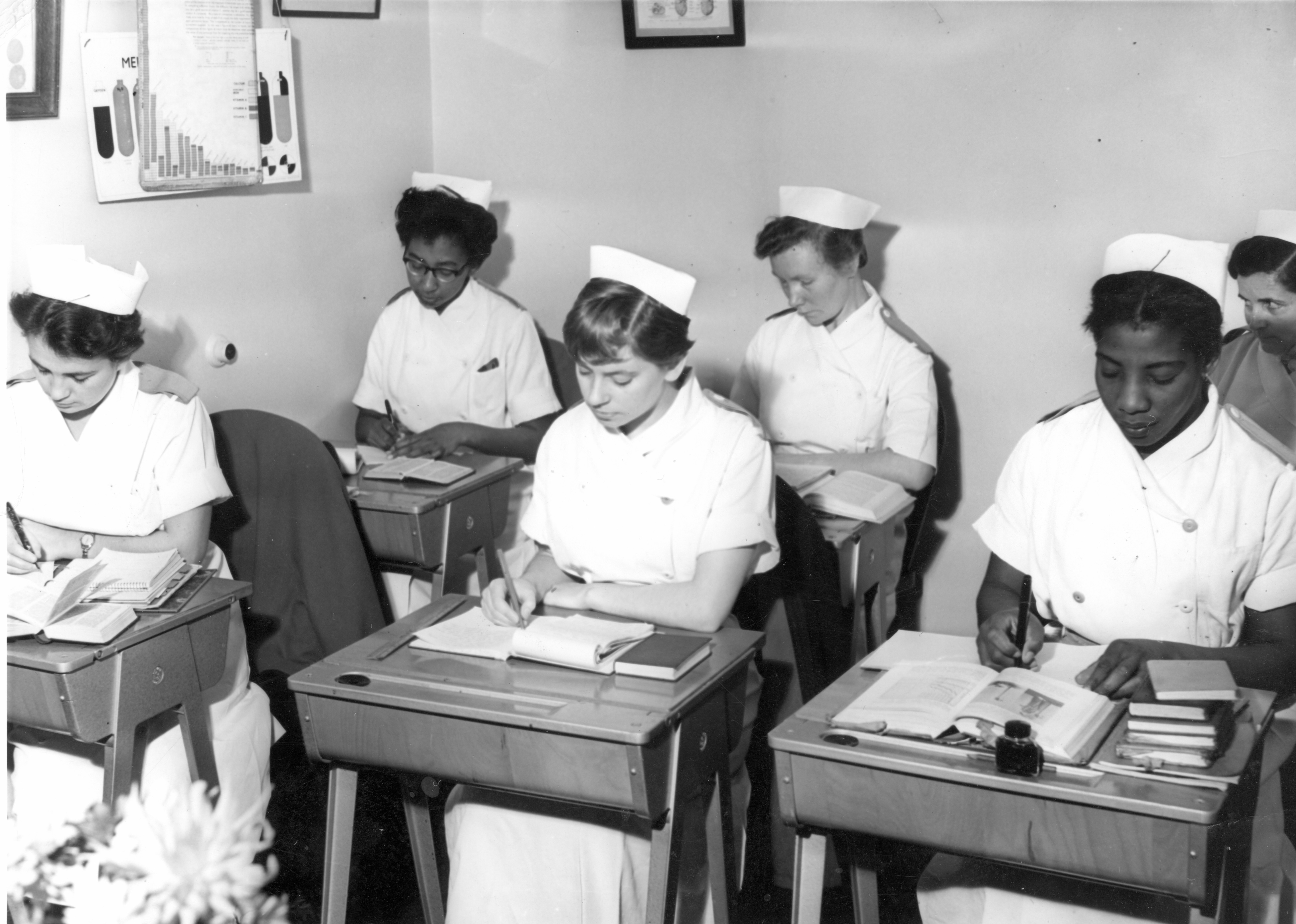 Student nurses in classroom c.1950.  black and white photograph.  Six young women wearing white uniforms and caps, sat at desks with textbooks open, taking notes. Posters on the wall and ink bottles on the desks.  Two of the women are black.  The original image was from a West London photographer and is likely from the Ealing / Acton area.