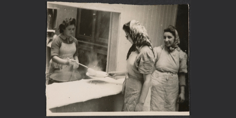 Greyscale image of a woman wearing a dress and apron serving food into a bowl from behind a counter. Another woman facing away from the camera wearing a dress, apron, headscarf and glasses is holding the bowl. Standing next to her is another woman facing the camera also wearing a dress, apron and headscarf.