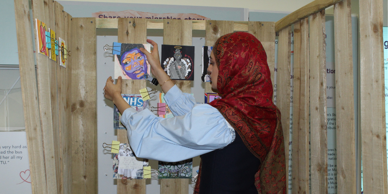 A brown woman in a red headscarf, light blue shirt and dark blue waistcoat glues and secures colourful, square artwork on wooden slats. In the background, parts of the surrounding exhibition on nursing and migration are visible.