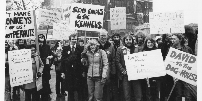 Black and white photograph from the Nursing Standard. It shows a group of people holding placards as part of the RCN's 'Pay Not Peanuts' campaign of 1979.