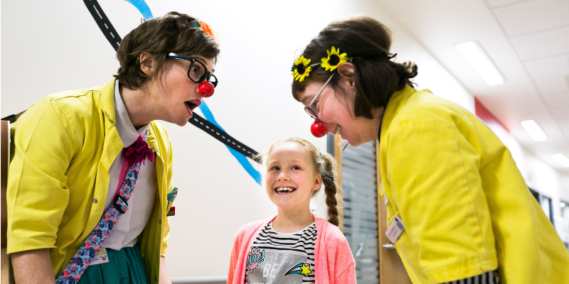Two white women dressed as healthcare clowns, wearing yellow jackets and red noses, interact joyfully with a smiling girl in a hospital hallway.