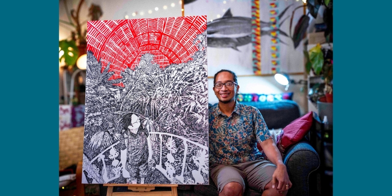 Yayen, a Filipino man with glasses and a blue floral shirt sits on a dark blue sofa beside a large, red and black artwork that he created. The room looks cosy, with plants and soft lighting. The drawing depicts a person in a leafy space under a red ceiling. 