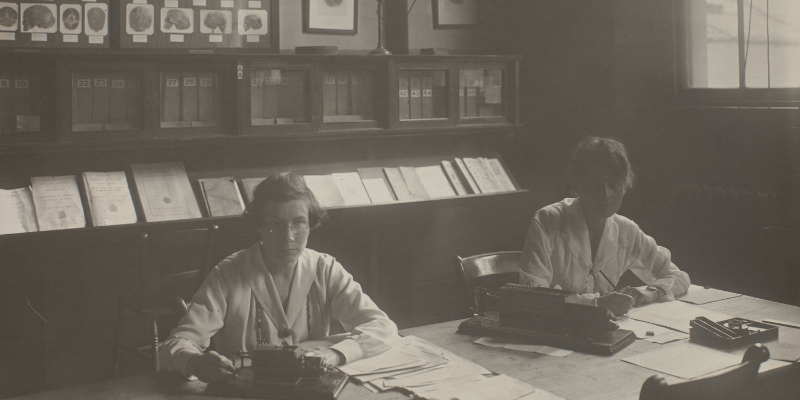 Greyscale image of two women working at a table in the Galton Laboratory. They have papers and equipment in front of them and there are shelves and books lining the wall behind them.