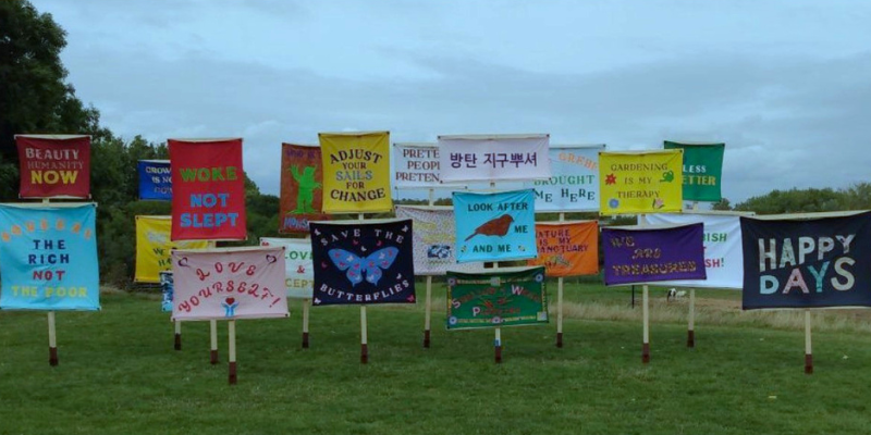 22 colourful, fabric banners, supported by wooden posts, with many different uplifting and motivational messages are displayed outdoors on a grassy area. Some of the messages include "Love Yourself", "Happy Days", "Adjust your sails for change", Gardening is my therapy" and "Woke not slept". Trees and the light grey, cloudy sky are visible in the background. 